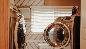 A laundry area with two front-loading washing machines, a white laundry basket, and a window with blinds.