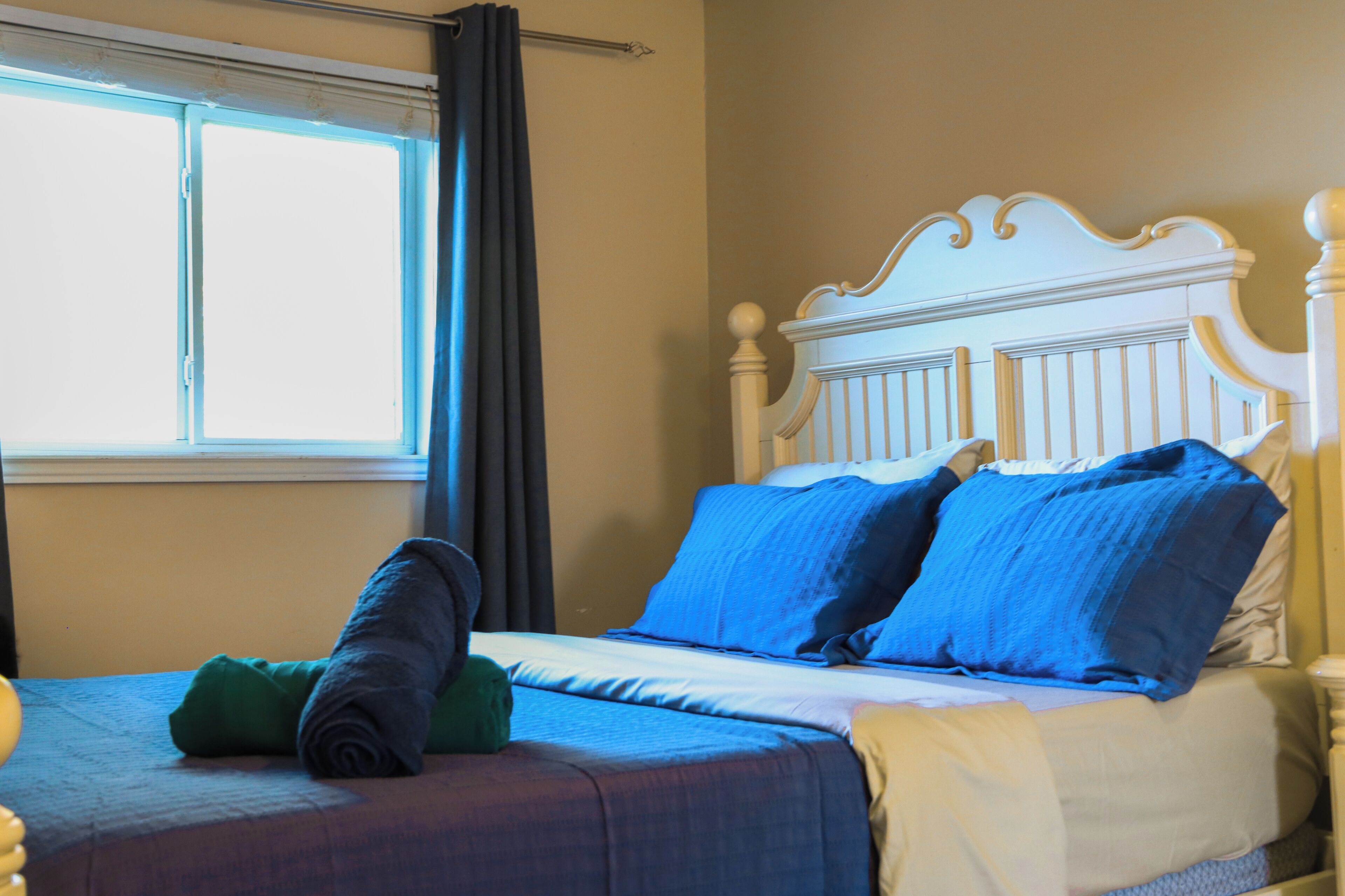 Un lit avec des oreillers bleus et verts, une tête de lit blanche et une fenêtre avec des rideaux.
