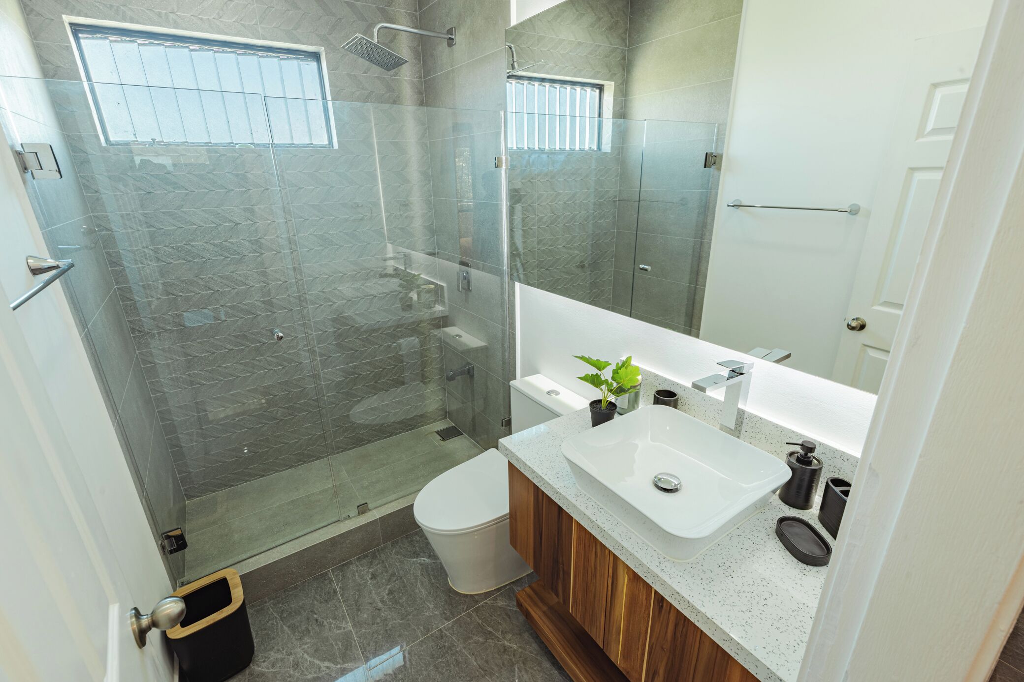 A modern bathroom with a glass-enclosed shower, a white toilet, a wooden vanity with a white sink, and a large mirror.