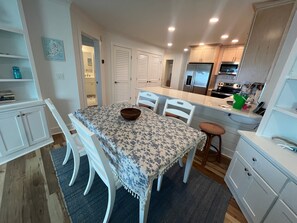 A dining area with a table set for two, white chairs, a bowl on the table, and a kitchen in the background with cabinets and appliances. - Stunning Oceanfront 2BR Villa with Panoramic Atlantic Views (Hilton Head Island)