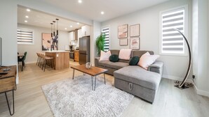A modern living room with a grey sofa, a wooden coffee table, and a white shag rug. There is a kitchen in the background with a refrigerator and a dining area with chairs. - ExoticLUSHquietRetreatKingBed|WIFI|Parking|WEM (Edmonton)