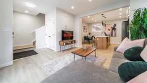 A modern living room with a sofa, coffee table, and a TV displaying 'NETFLIX'. There is a staircase leading to another room, a dining area, and a kitchen visible in the background. - ExoticLUSHquietRetreatKingBed|WIFI|Parking|WEM (Edmonton)