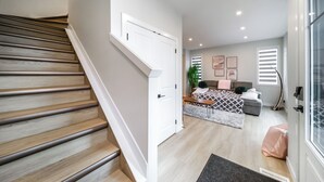A modern interior with a wooden staircase, a bed with patterned bedding, a grey sofa, and a large window with blinds. - ExoticLUSHquietRetreatKingBed|WIFI|Parking|WEM (Edmonton)