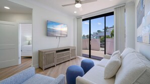 A modern living room with a large window, a ceiling fan, and a flat-screen TV mounted on the wall. - Surfside G19 - Condo in Surfside Resort with Many Shared Amenities (Miramar Beach)