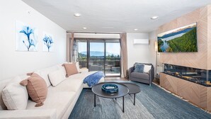 A modern living room with a sofa, a coffee table, and a fireplace. There is a large window with a view of mountains and a lake. - Newley renovated ski-in/ ski-out duel condo w communal hot tub and great views (Whitefish)