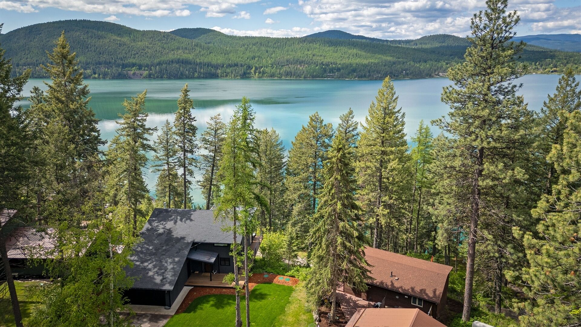 A lakeside property with multiple buildings, surrounded by dense forest and mountains in the background.