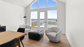 A modern room with a wooden floor, white walls, and a large window offering a view of the beach. There is a wooden table, two white chairs, and a black floor lamp. - Foley’s Harbour House (Alberton)