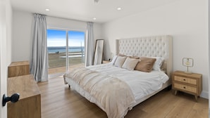 A bedroom with a large bed, a nightstand, a wooden dresser, and a view of the beach through a sliding glass door. - Foley’s Harbour House (Alberton)