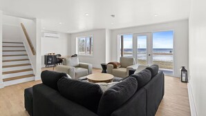 A spacious living room with a large black sofa, a round coffee table, and a beige armchair. A staircase with wooden steps leads to a dining area with a small table and chairs. Large windows offer a view of the beach and ocean. - Foley’s Harbour House (Alberton)