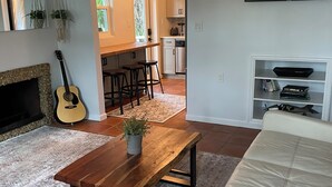 Ein Wohnzimmer mit Couch, Kamin, Holztisch und Gitarre, im Hintergrund eine Küche.