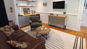 Ein Wohnzimmer mit braunem Ledersofa, einem Couchtisch, einem an der Wand montierten Fernseher und einer Küchenzeile mit Arbeitsplatte und Schränken.
