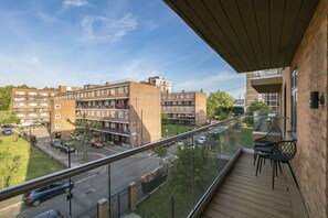 Un balcón con vista a un edificio de varios pisos, una calle con coches y un área verde.