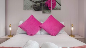 Un lit avec une literie blanche et deux oreillers roses, une table de chevet avec une lampe, et un mur orné d’une œuvre d’art représentant des roses.