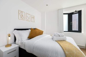Une chambre à coucher avec un lit, du linge de lit blanc, une lampe dorée, une fenêtre donnant sur un beau paysage et des œuvres d’art mural.