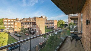 Un balcon donnant sur un immeuble résidentiel à plusieurs étages, une rue animée et un ciel dégagé.