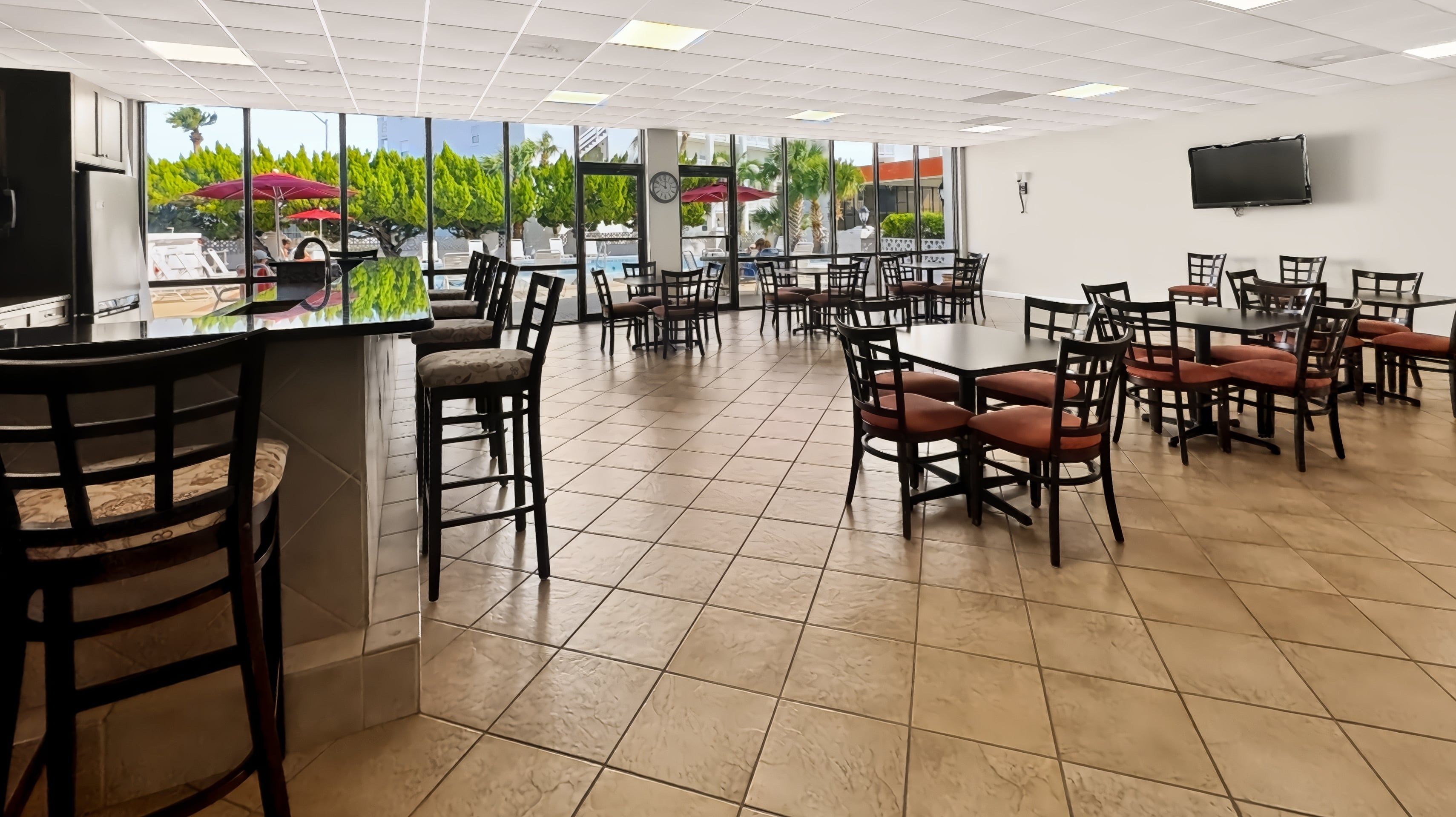 A spacious indoor area with multiple tables and chairs, large windows offering views of a pool and greenery, and a flat-screen TV mounted on the wall.