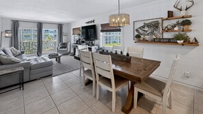 A modern living room with a dining area, large windows, and a kitchen in the background.