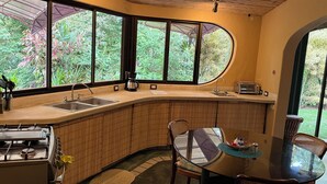 Una cocina con estufa a gas, lavamanos y cafetera. Hay una mesa redonda con sillas y vistas al exterior, con vegetación.