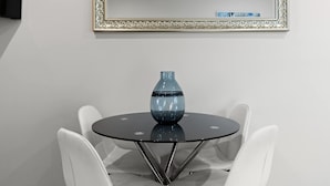 Une table ronde en verre, surmontée d’un vase bleu, entourée de chaises blanches, le tout placé sous un miroir décoratif.
