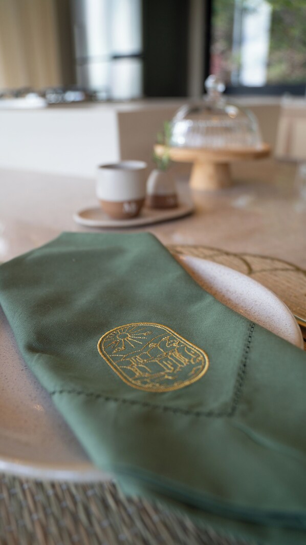 Una mesa con mantel verde y un escudo bordado, una taza blanca y un recipiente de vidrio.