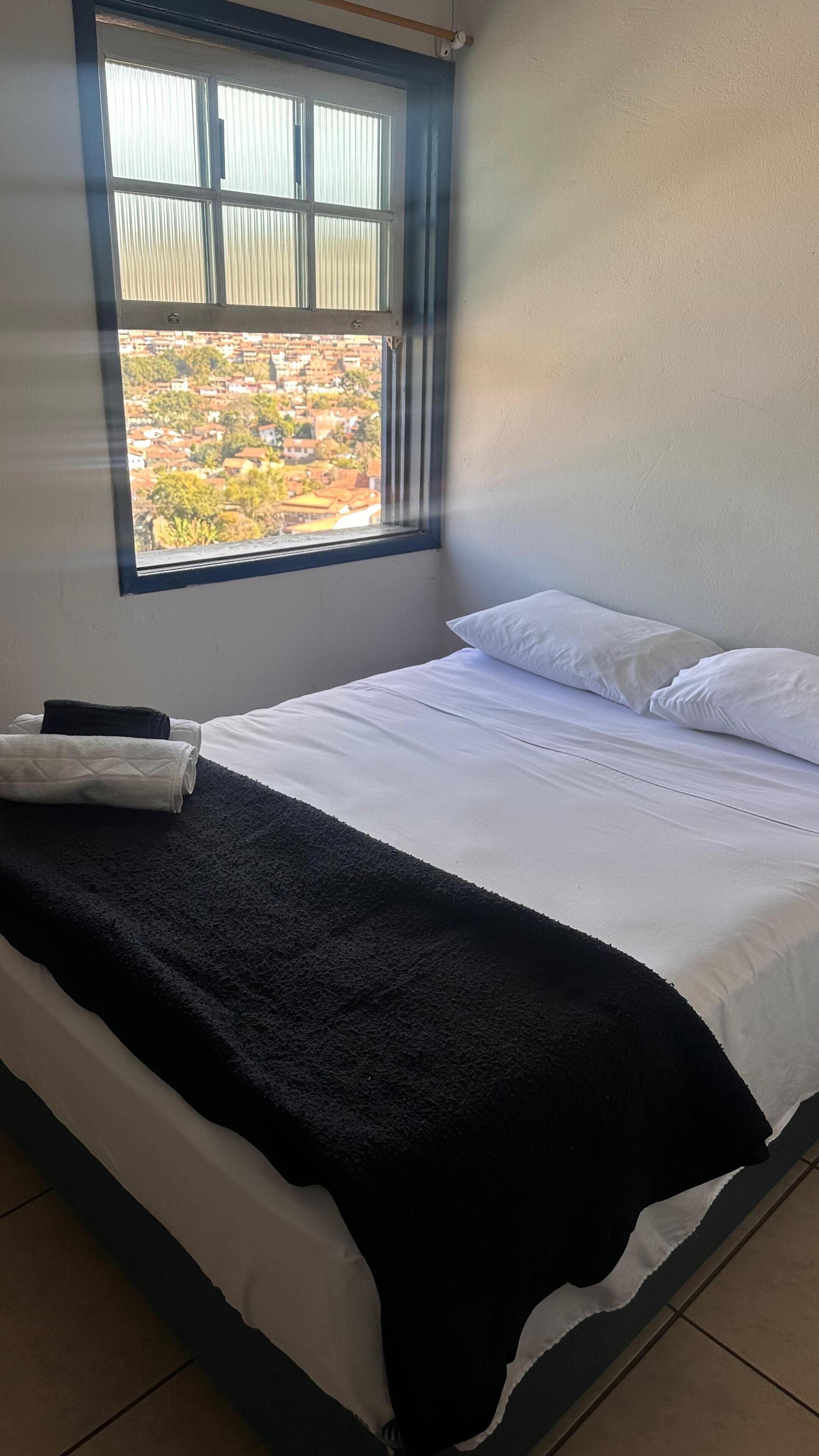 A neatly made bed with white linens and a black throw blanket, a window with blinds, and a view of a residential area.