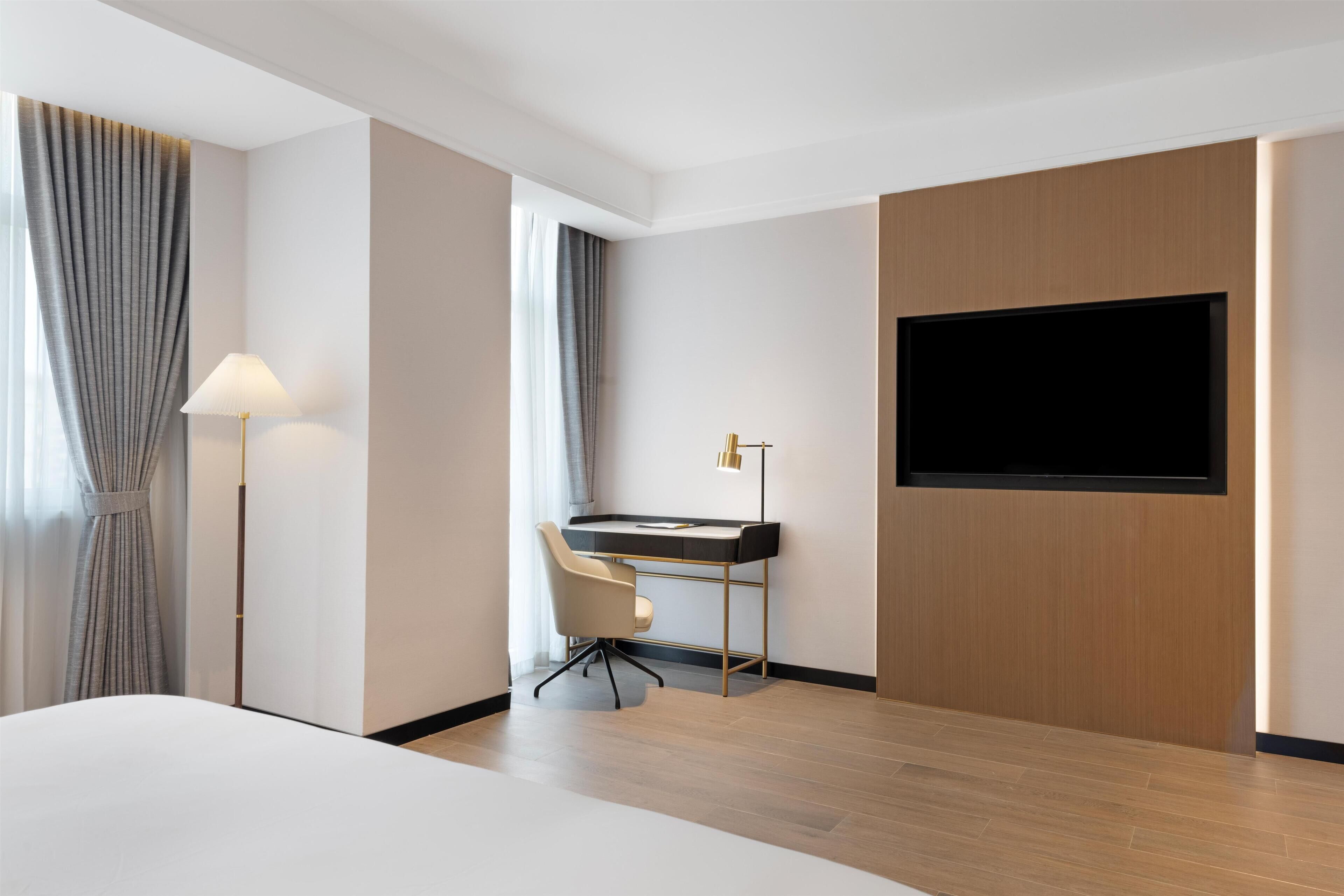A modern hotel room with a bed, a desk with a chair, a flat-screen TV mounted on the wall, and a lamp.
