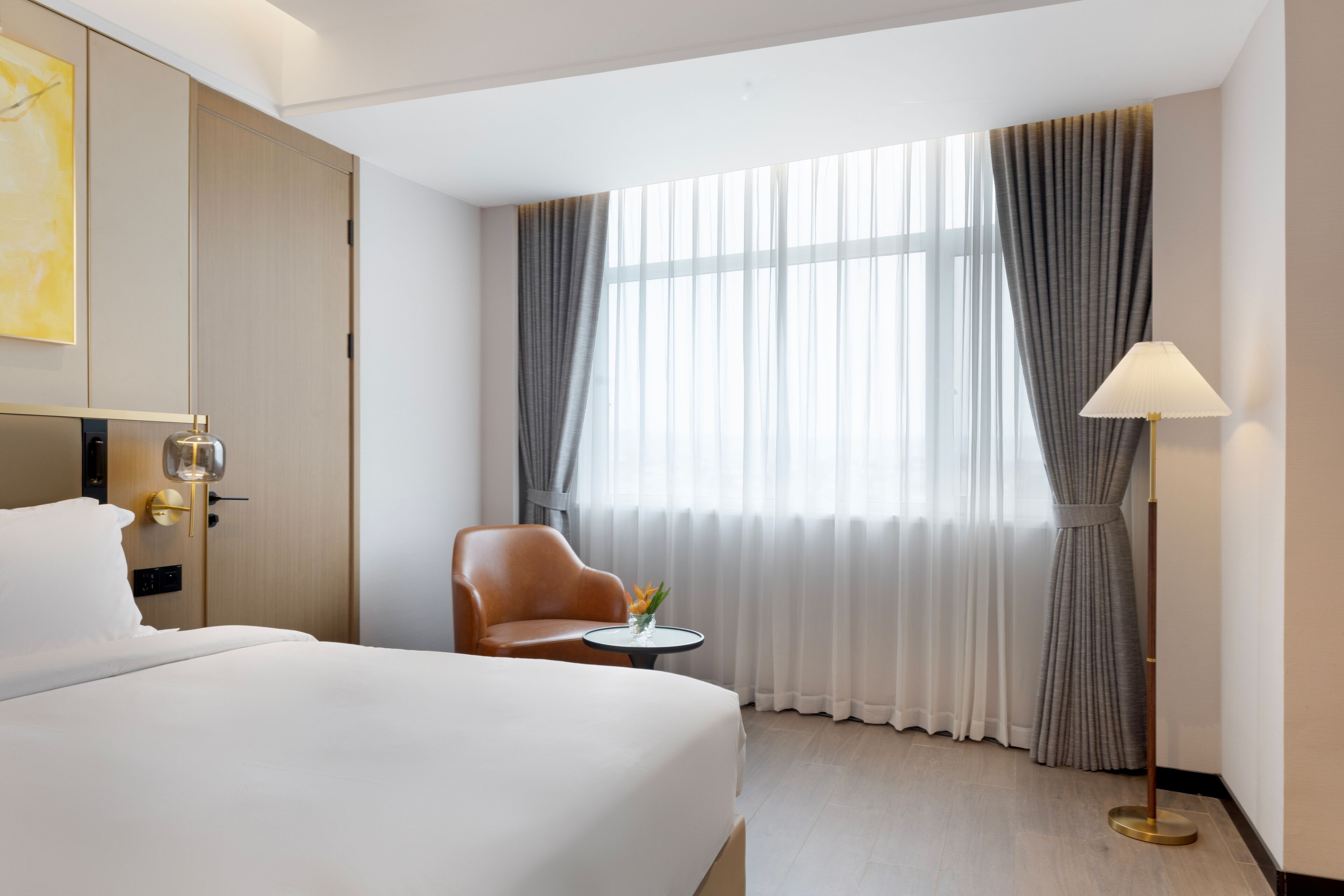 Quarto de hotel com uma cama grande, uma cadeira, uma mesinha com um vaso e um abajur.
