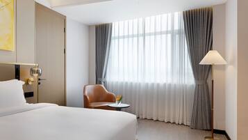 Quarto de hotel com uma cama grande, uma cadeira, uma mesinha com um vaso e um abajur.