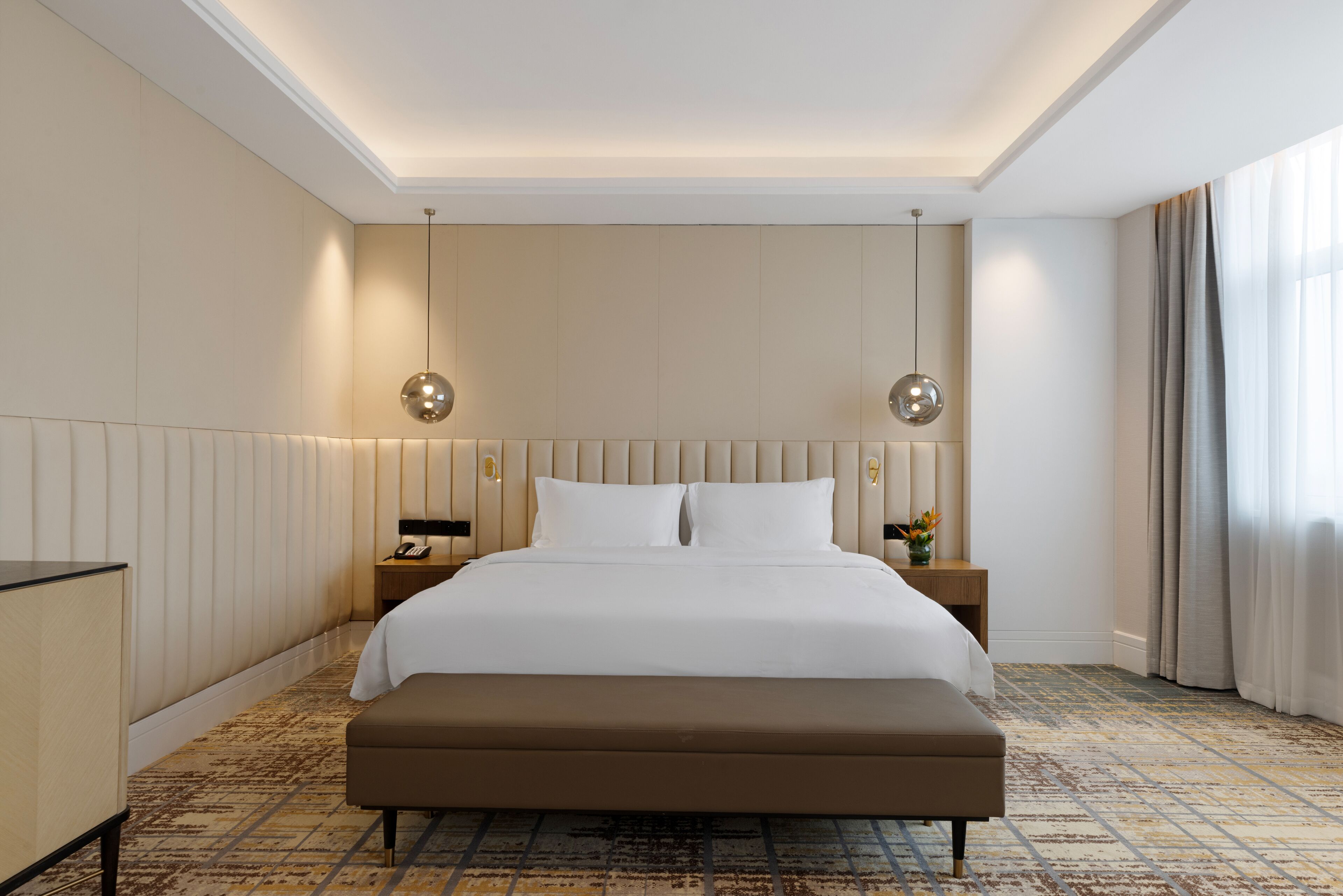 Habitación de hotel con una cama grande, un banco, dos mesitas de noche y una mesita con lámpara.