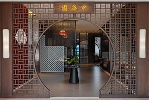A traditional Chinese gate with a sign reading "Garden Center". Inside, there is a round table with a potted plant and a modern interior with wooden partitions and a visible staircase. - Golden Tulip Kin-Oasis Kinshasa (Kinshasa)