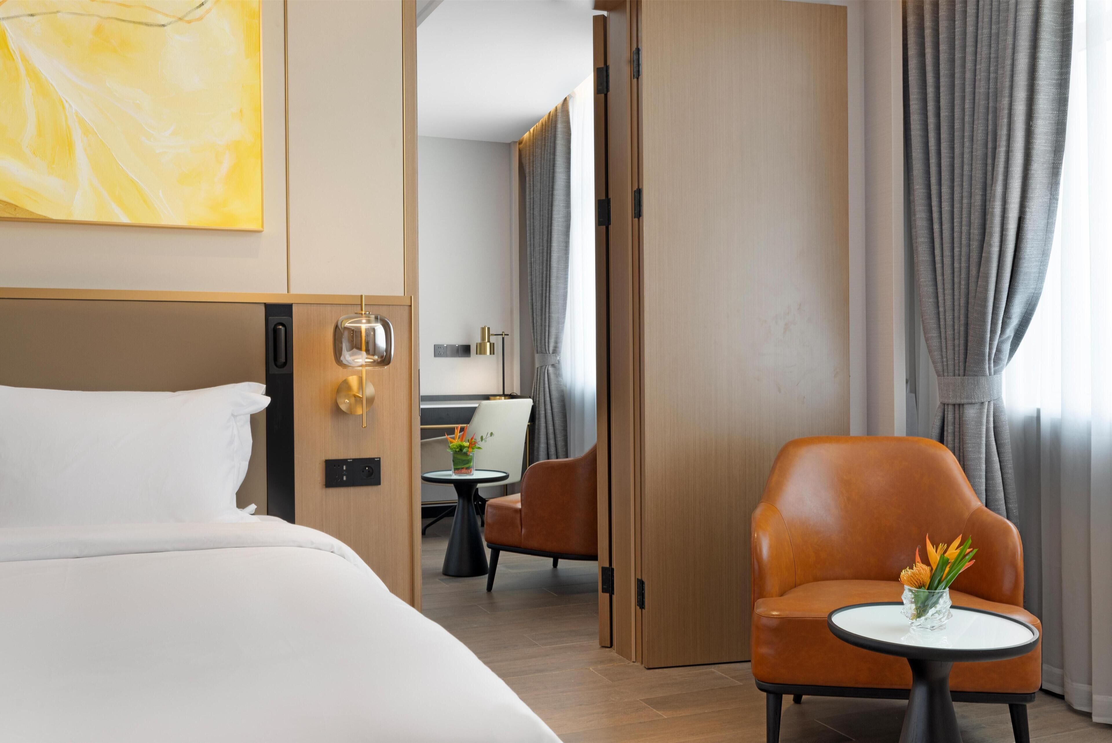 Una habitación de hotel con cama, una silla, una mesita con un jarrón de flores y una puerta que da a otra habitación.