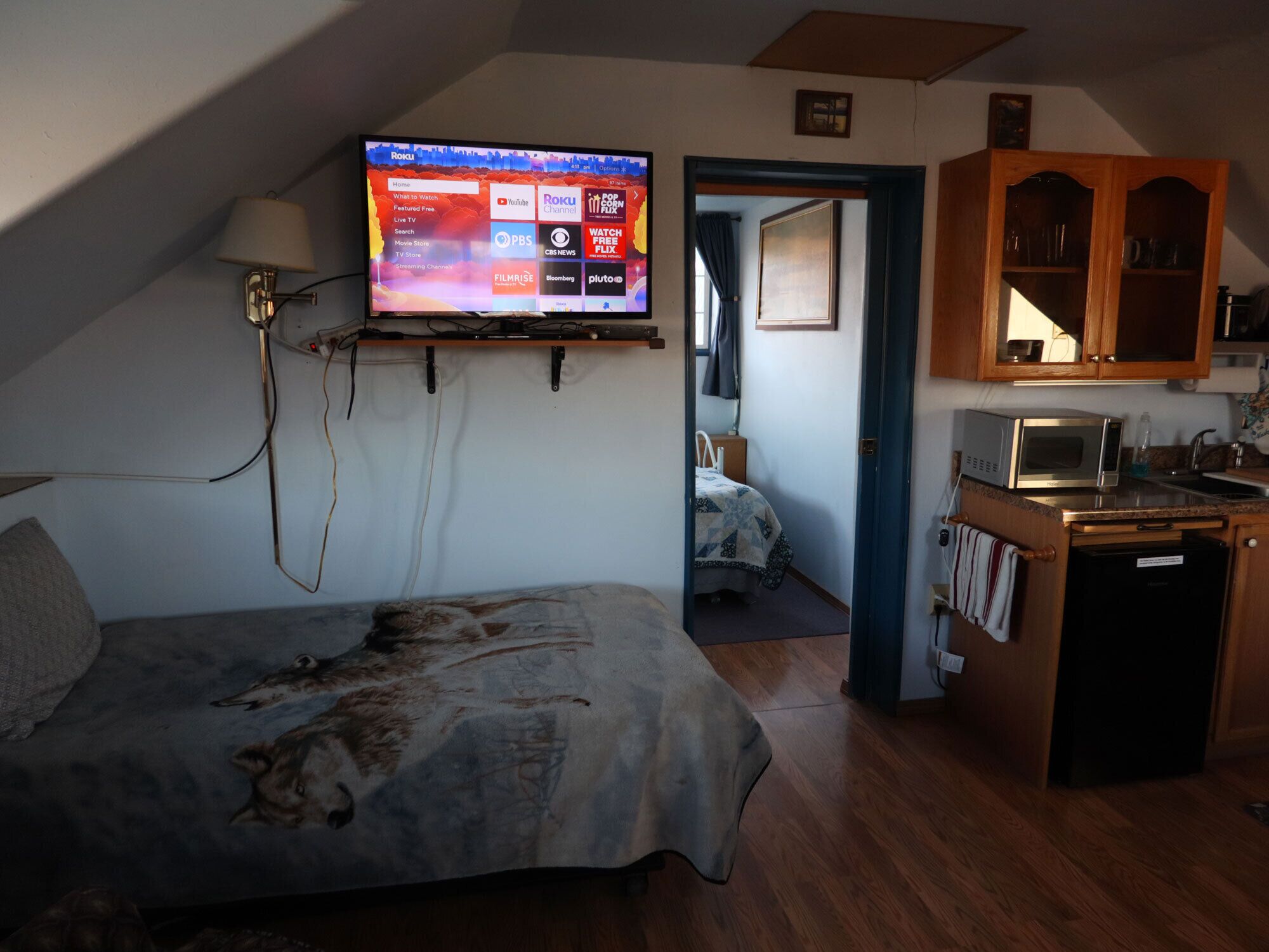 A bedroom with a bed, a TV mounted on the wall, a wooden cabinet, and a kitchenette area.