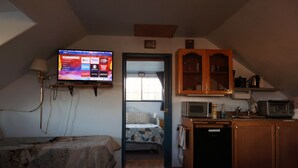 Ein kleines Zimmer mit einem Bett, einem an der Wand befestigten Fernseher, einer Küchenzeile und einer Tür zu einem weiteren Raum.