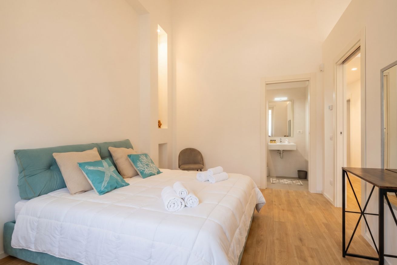 Ein Schlafzimmer mit Bett, Kissen, einem Sessel, einem Tisch und einem Badezimmer im Hintergrund.