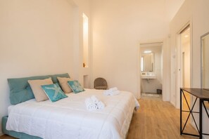 Quarto com cama, almofadas, uma cadeira, uma mesa e um banheiro visível ao fundo.