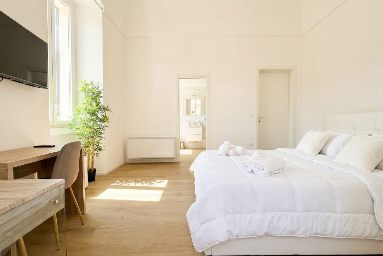 A bedroom with a bed, a desk, a chair, a potted plant, and a TV.