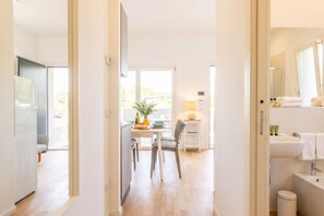 A modern hotel room with a wooden floor, a dining area with a table and chairs, a kitchen area, and a bathroom with a sink and mirror.