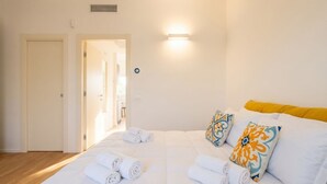 Une chambre d’hôtel avec un lit, des serviettes pliées, une table de chevet, une lampe fixée au mur et une porte donnant sur une autre pièce.