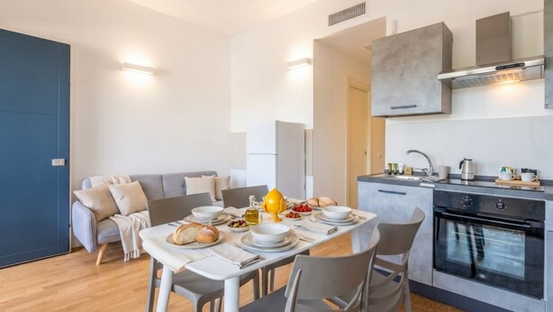 Une cuisine moderne dotée d’un four encastré, un coin repas avec une table préparée pour deux personnes, et un canapé gris en arrière-plan.