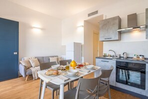 A modern kitchen with a built-in oven, a dining area with a table set for two, and a grey sofa in the background.