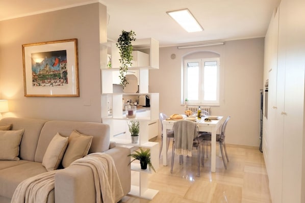 A modern living room with a sofa, a dining area with a table and chairs, and a kitchen area with cabinets and a hanging plant.