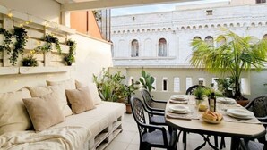 A cozy outdoor seating area with a sofa and dining table, surrounded by potted plants and overlooking a historic building with arches.