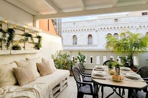 A cozy outdoor seating area with a sofa and dining table, surrounded by potted plants and overlooking a historic building with arches.