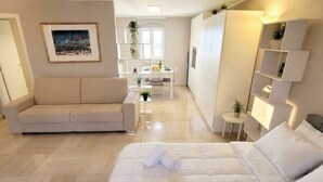 A modern hotel room with a large bed, a sofa, a dining area, and a kitchen.