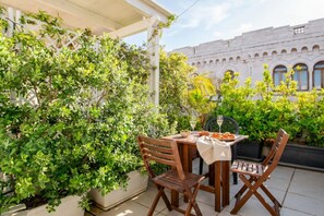 An outdoor patio with a table set for two, surrounded by lush greenery and a white building in the background.