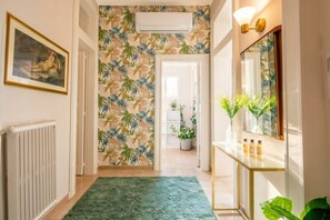 A hallway with a patterned wallpaper, a radiator, a green rug, a mirror, and a plant.