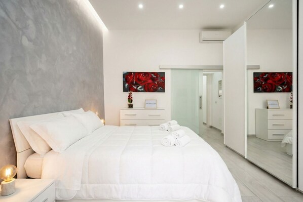 A modern bedroom with a large bed, a bedside table with a lamp, a dresser, and a mirror.