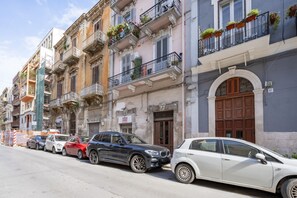 A row of parked cars along a street with multi-story buildings featuring balconies with potted plants.