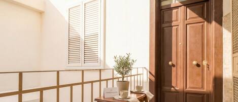 A balcony with a wooden railing, a potted plant, a table with a sign, and a brown double door.