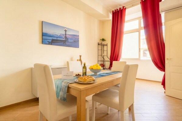 A dining area with a wooden table, white chairs, a painting on the wall, a bowl of fruit, and a bottle of olive oil.